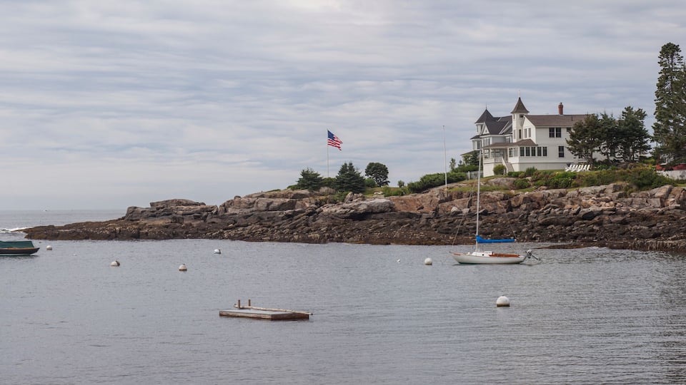 A coastal scene with a lighthouse, rocky shoreline, and a large white house with a flag.