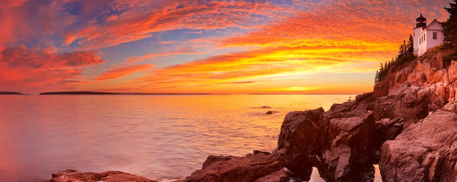 Un atardecer costero con un faro, costa rocosa y mar en calma.