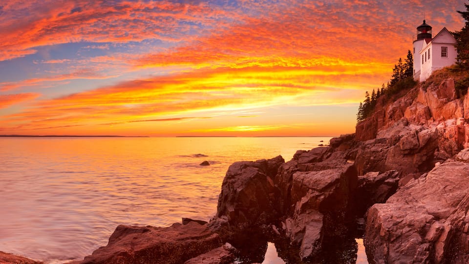 A coastal sunset with a lighthouse, rocky shoreline, and calm sea.