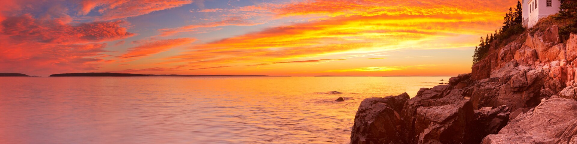 Un atardecer costero con un faro, costa rocosa y mar en calma.