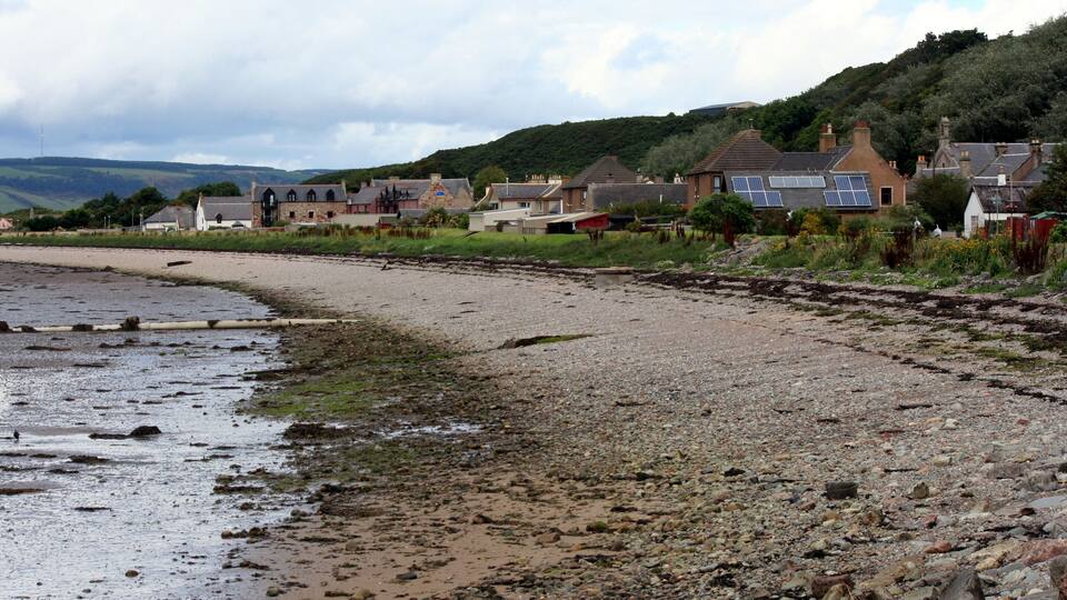 A pebbly beach with houses and a hill in the background.