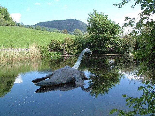 A large bird sculpture in a pond surrounded by greenery and hills.