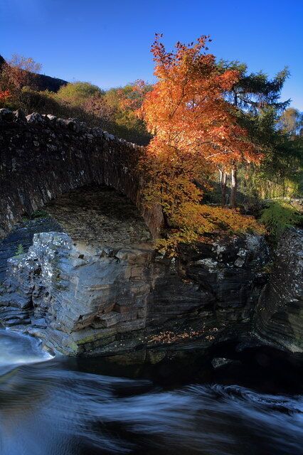 A stone bridge over a flowing river, surrounded by autumn foliage.