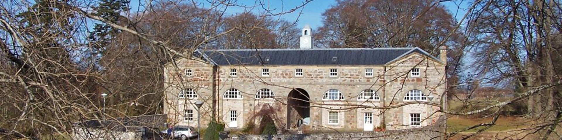 Un edificio de piedra histórico con un arco central, rodeado de árboles y un cielo azul despejado.