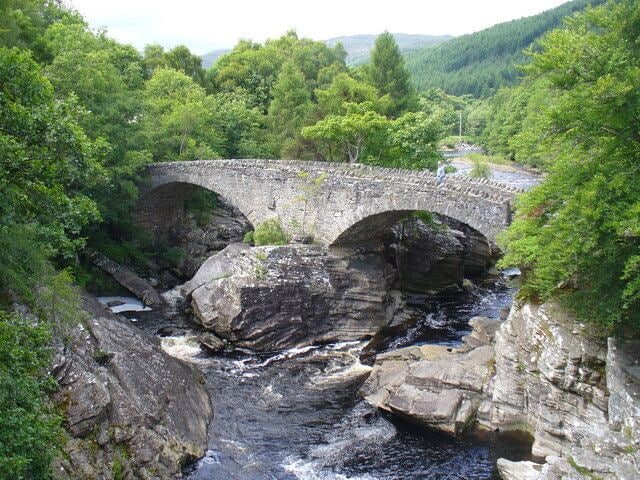 A stone bridge over a river, surrounded by lush greenery and rocky terrain.