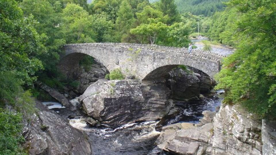 A stone bridge over a river, surrounded by lush greenery and rocky terrain.