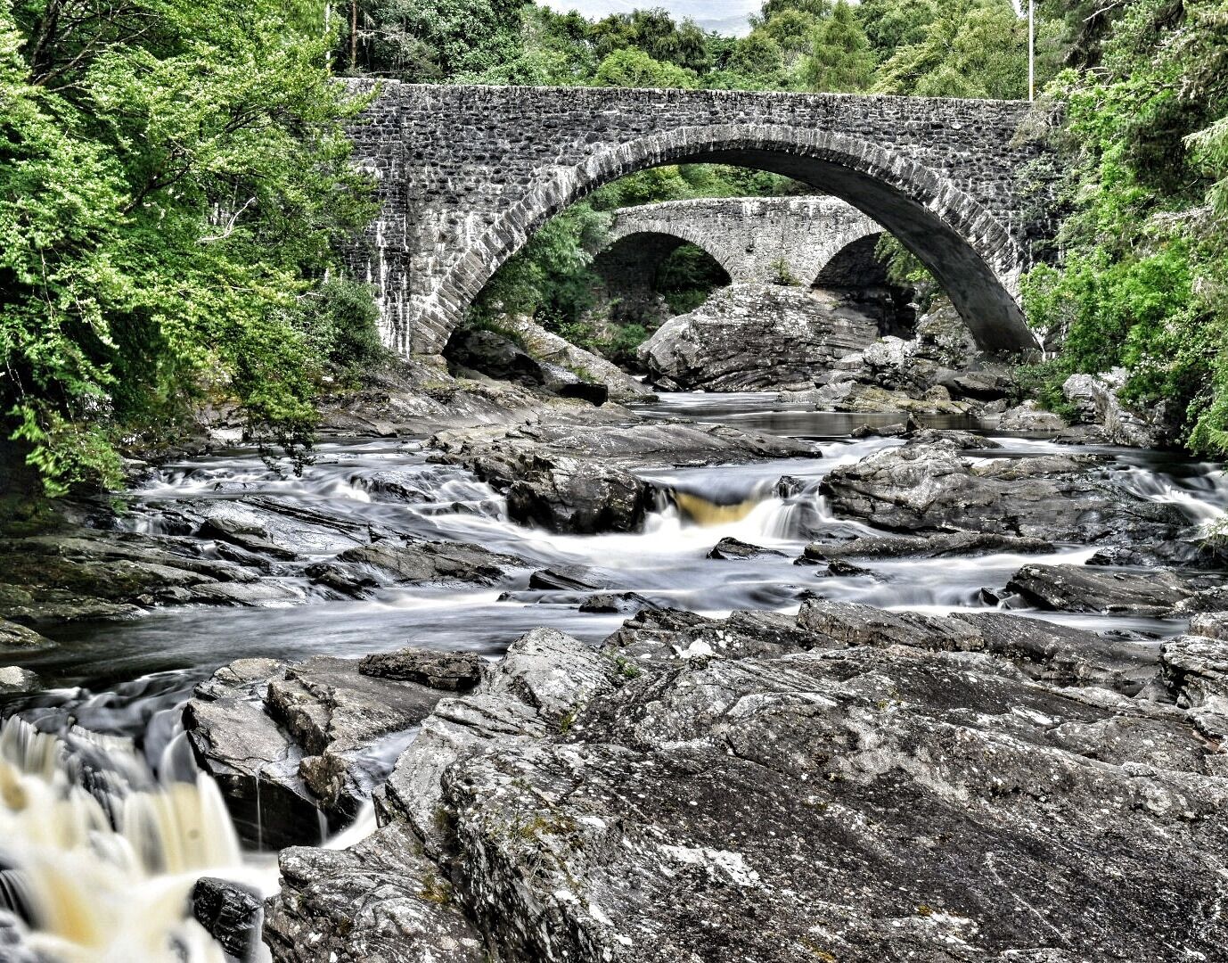 A stone bridge over a flowing river with surrounding greenery.