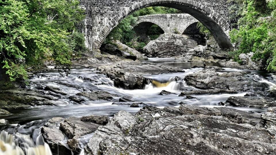 A stone bridge over a flowing river with surrounding greenery.