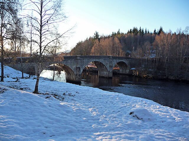 A stone bridge over a river with snow on the ground and trees in the background.