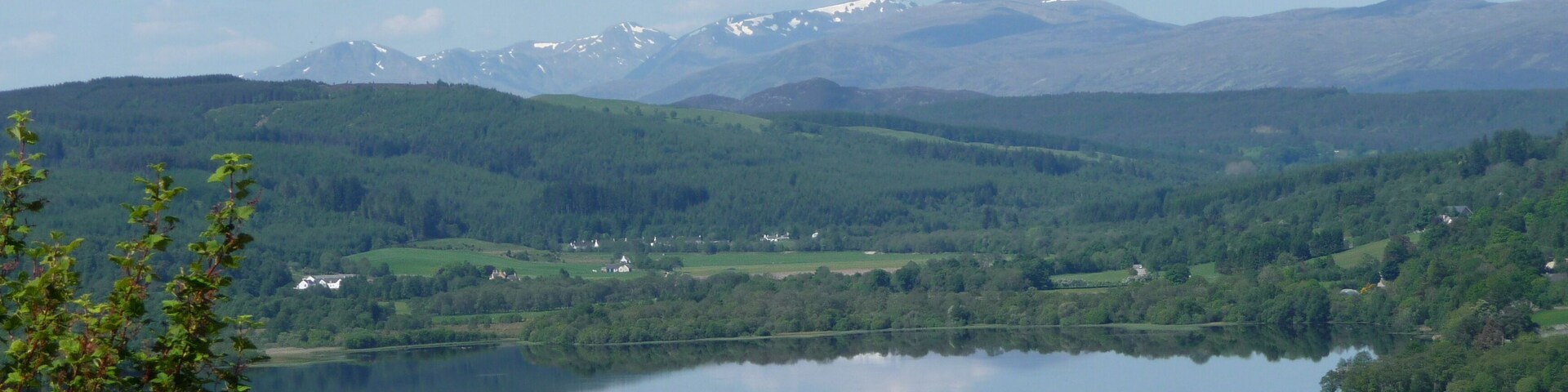 Um lago rodeado por colinas verdes e montanhas com picos nevados ao longe.