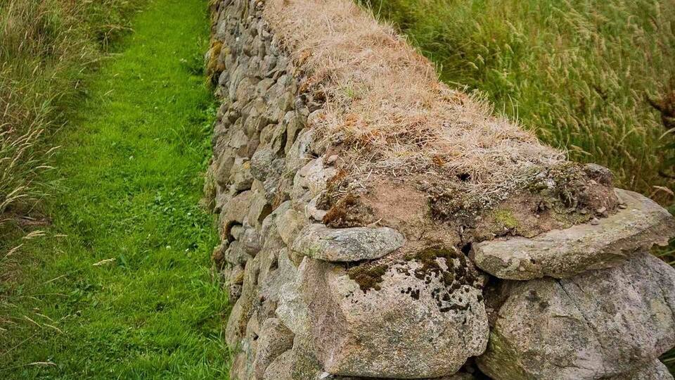 A stone wall beside a grassy path with fields and hills in the background.