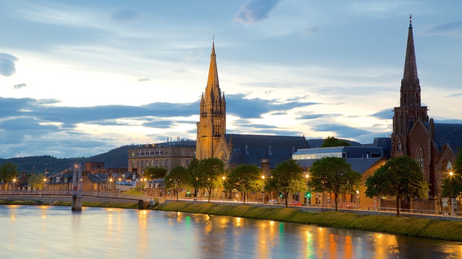 Un río con dos imponentes campanarios de iglesia, un puente y un paisaje urbano con edificios bajo un cielo crepuscular.
