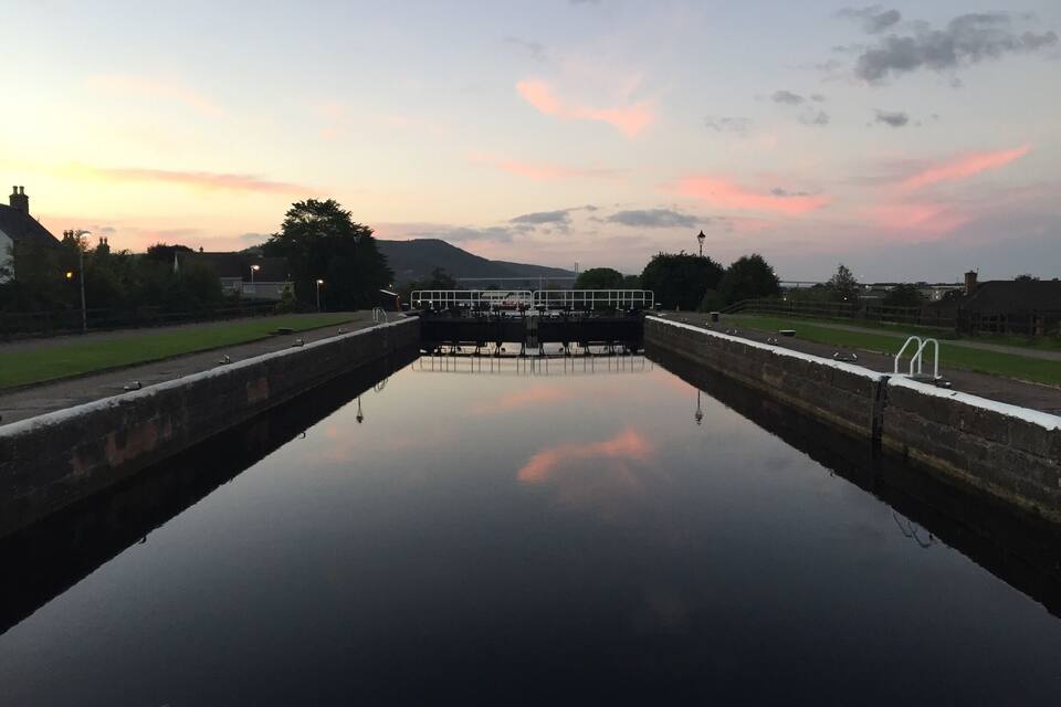 A calm canal with a lock gate, surrounded by greenery and buildings, under a twilight sky.