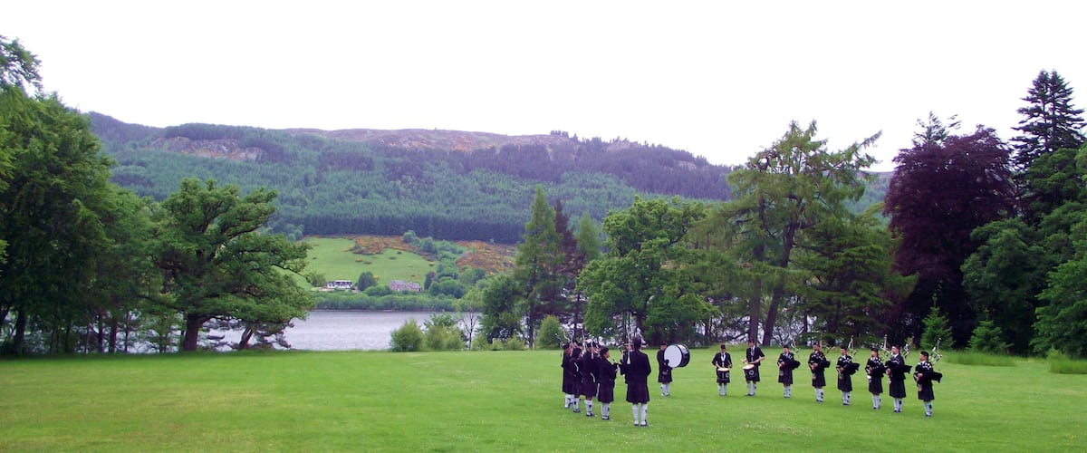 A group of people in traditional attire standing in a grassy field with a lake and hills in the background.