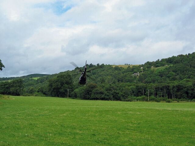 Um helicóptero sobrevoando um campo verde com uma colina arborizada ao fundo.