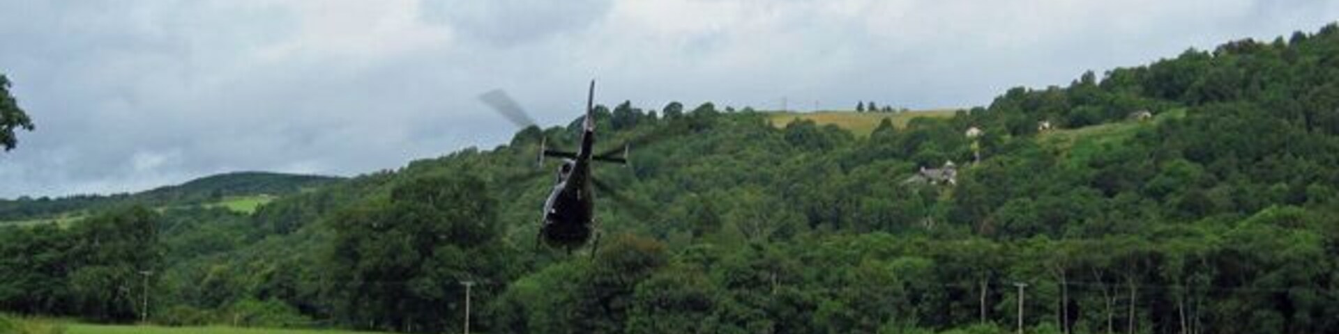 Um helicóptero sobrevoando um campo verde com uma colina arborizada ao fundo.