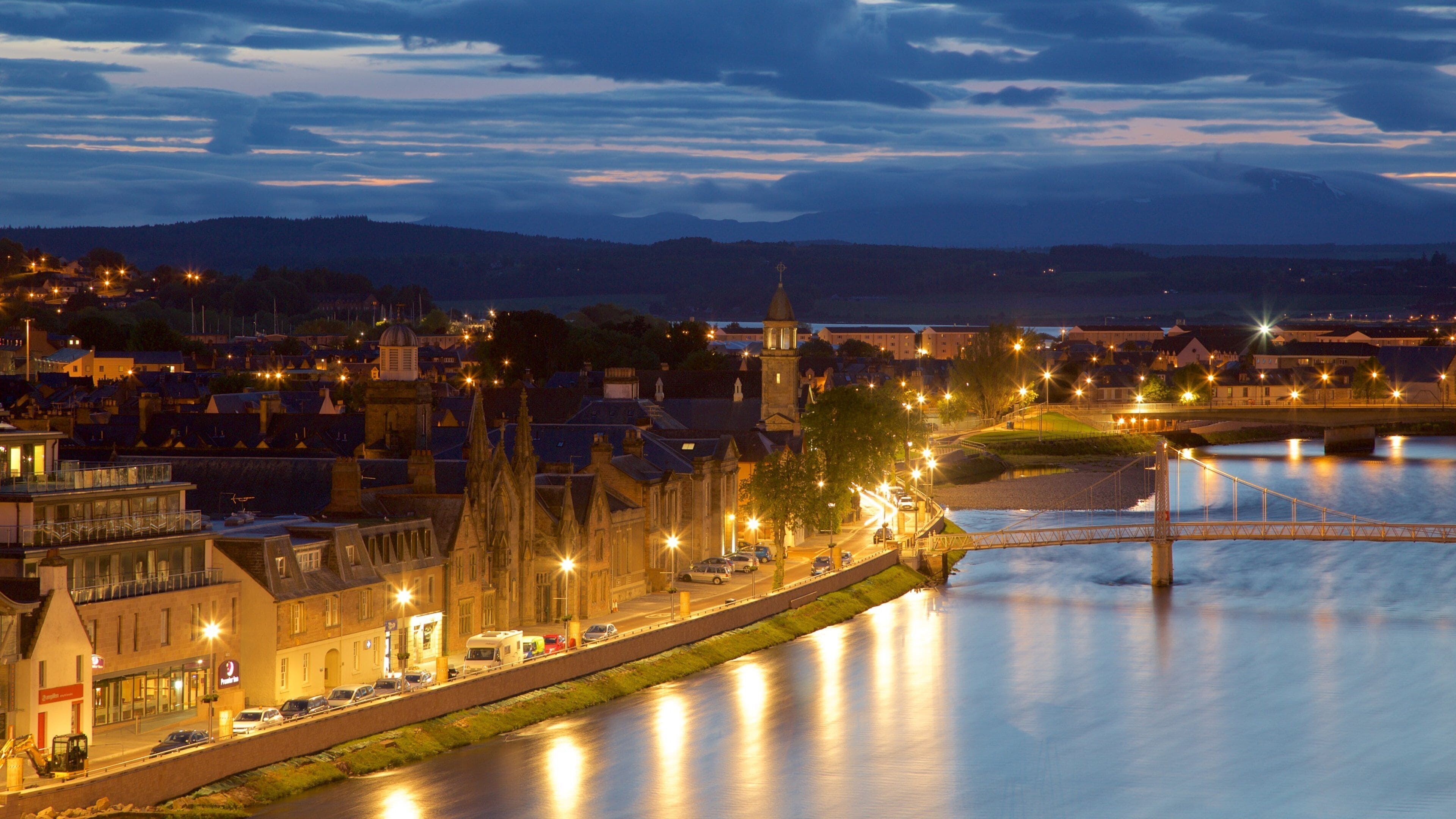 A riverside town at dusk with illuminated buildings, a prominent church tower, and a bridge over the river.