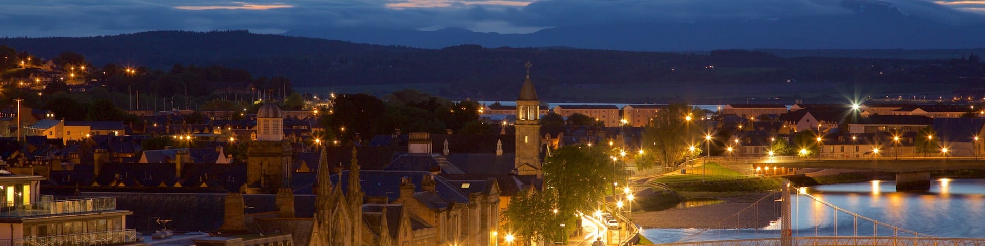A riverside town at dusk with illuminated buildings, a prominent church tower, and a bridge over the river.