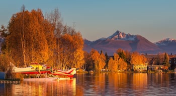 A lake with a red and white seaplane docked on a small island, surrounded by autumn-colored trees and a mountain range in the background.