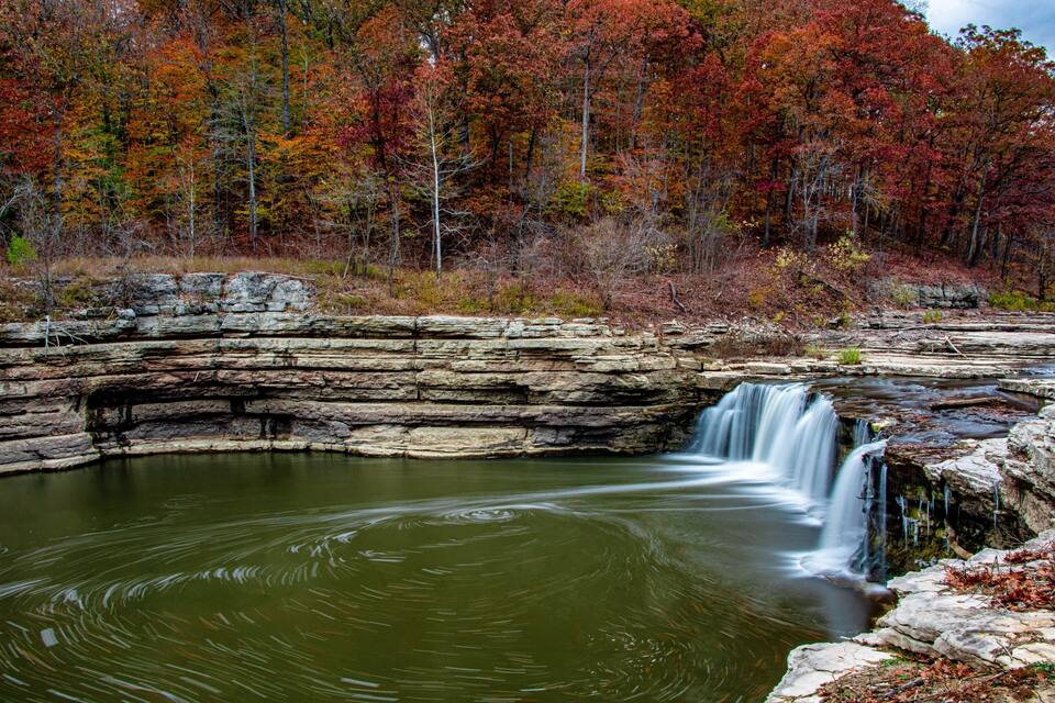 Une cascade d’eau se déversant sur un promontoire rocheux et se jetant dans un bassin paisible, le tout entouré d’arbres aux couleurs automnales.