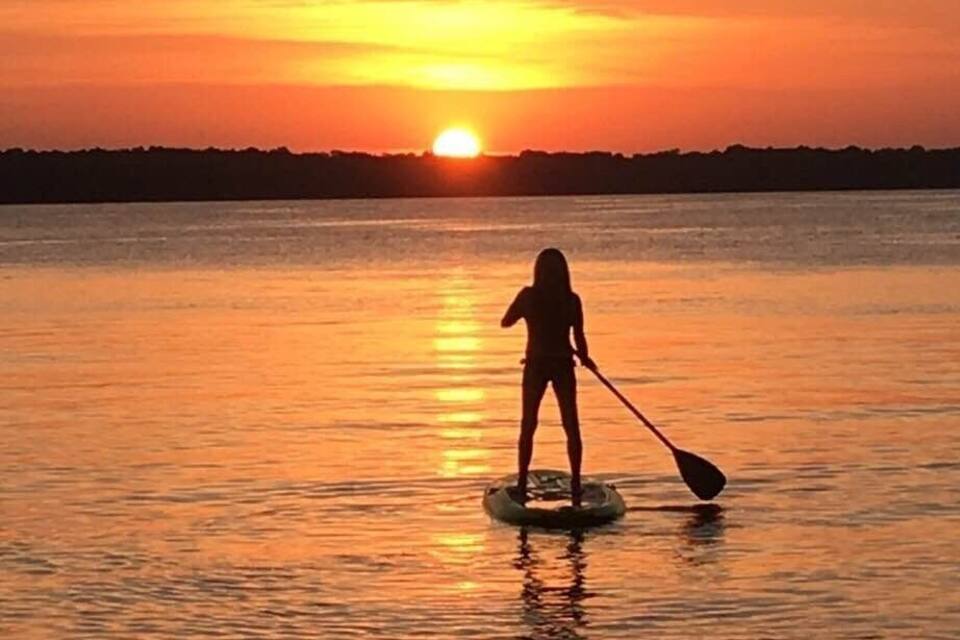 Una persona surfejant en una taula de pala al capvespre, amb el sol ponent-se a l'horitzó.