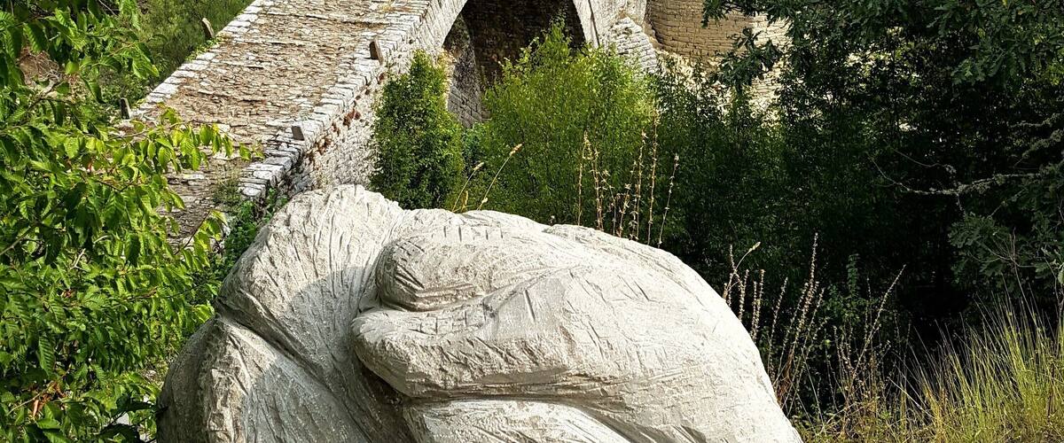 A stone bridge with arches, surrounded by lush greenery and a large rock sculpture.