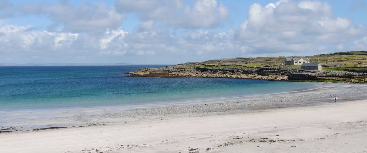 A beach with clear blue water, a rocky outcrop, and a small house on the shore.