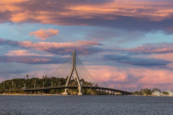 Uma ponte suspensa com montanhas ao fundo e um céu ao pôr do sol.