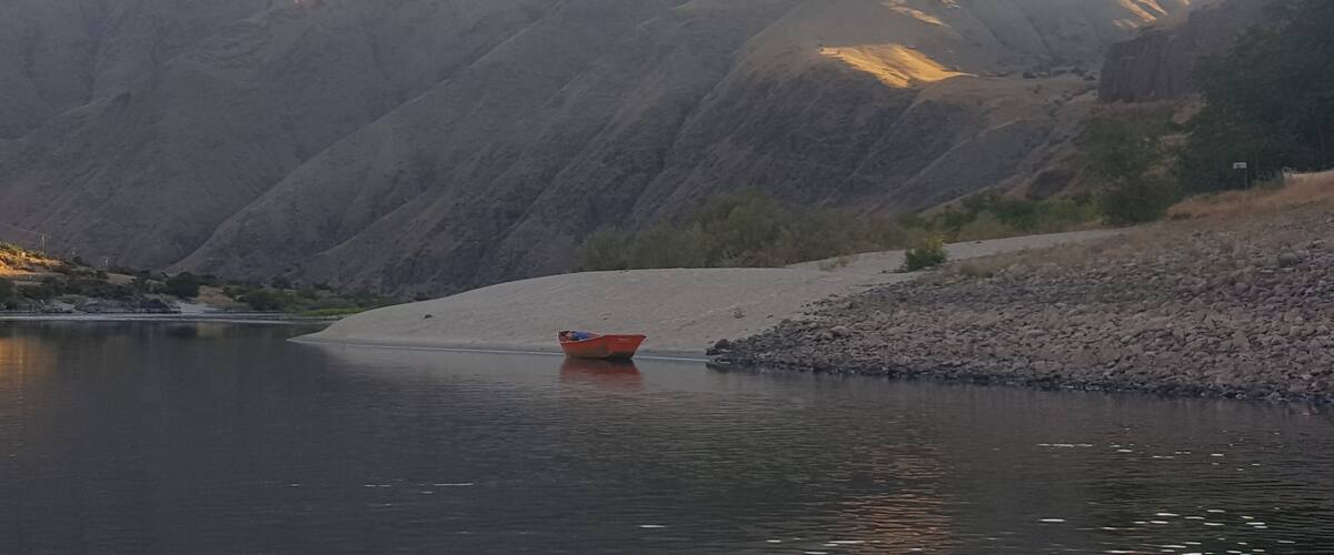 A calm lake with a small red boat in the foreground, surrounded by rocky terrain and mountains in the background.