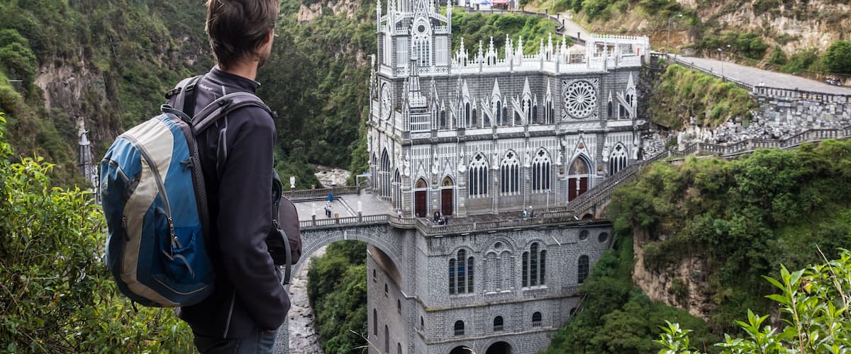 A person with a backpack overlooking a cathedral with intricate architecture, set against a lush green valley.