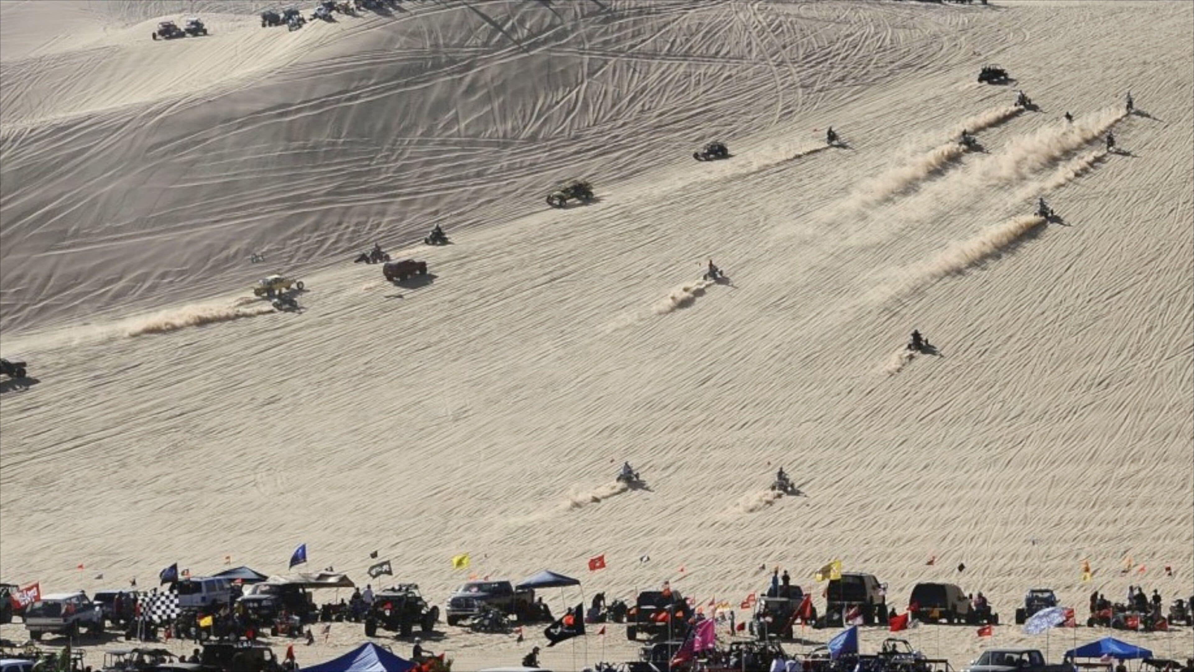 A snowy mountain with vehicles racing down the slope and spectators gathered at the bottom.