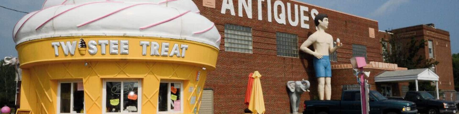 A yellow ice cream cone-shaped building with 'TW STEE TREAT' written on it, in front of a brick building labeled 'ANTIQUES'.