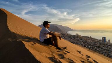 Young man sitting at the summit of enormours sand dune and overlooking city and ocean at the sunset