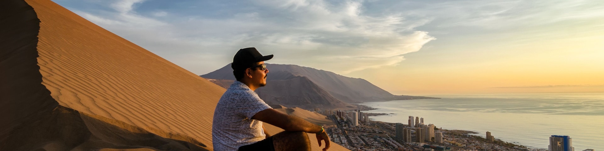 Young man sitting at the summit of enormours sand dune and overlooking city and ocean at the sunset