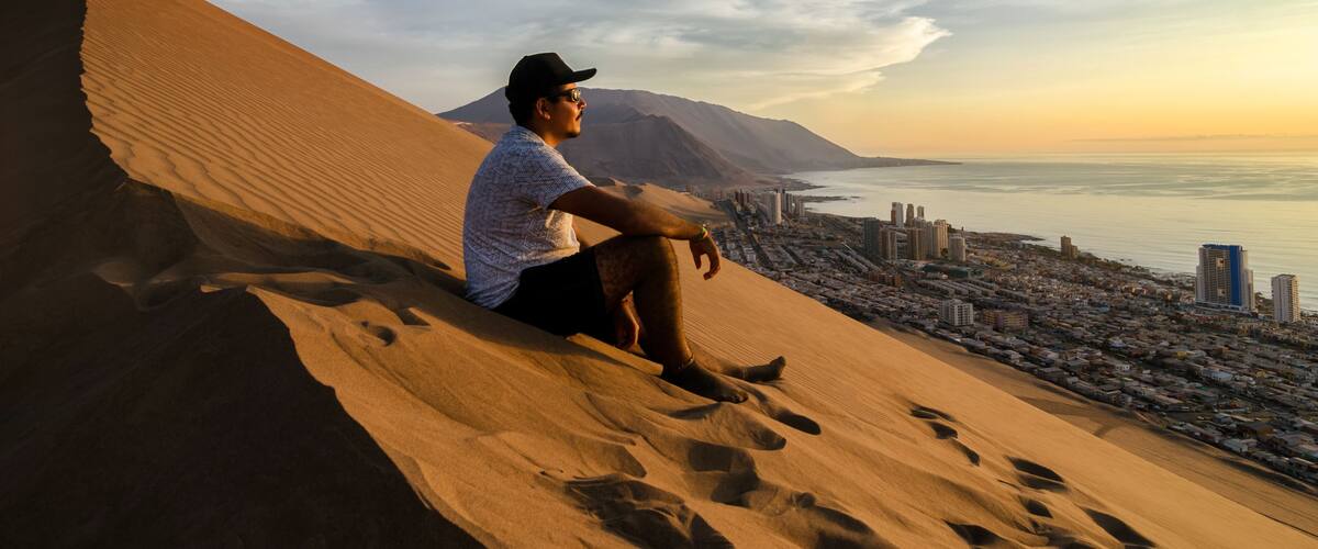 A person sitting on a sand dune overlooking a coastal cityscape with mountains in the background.