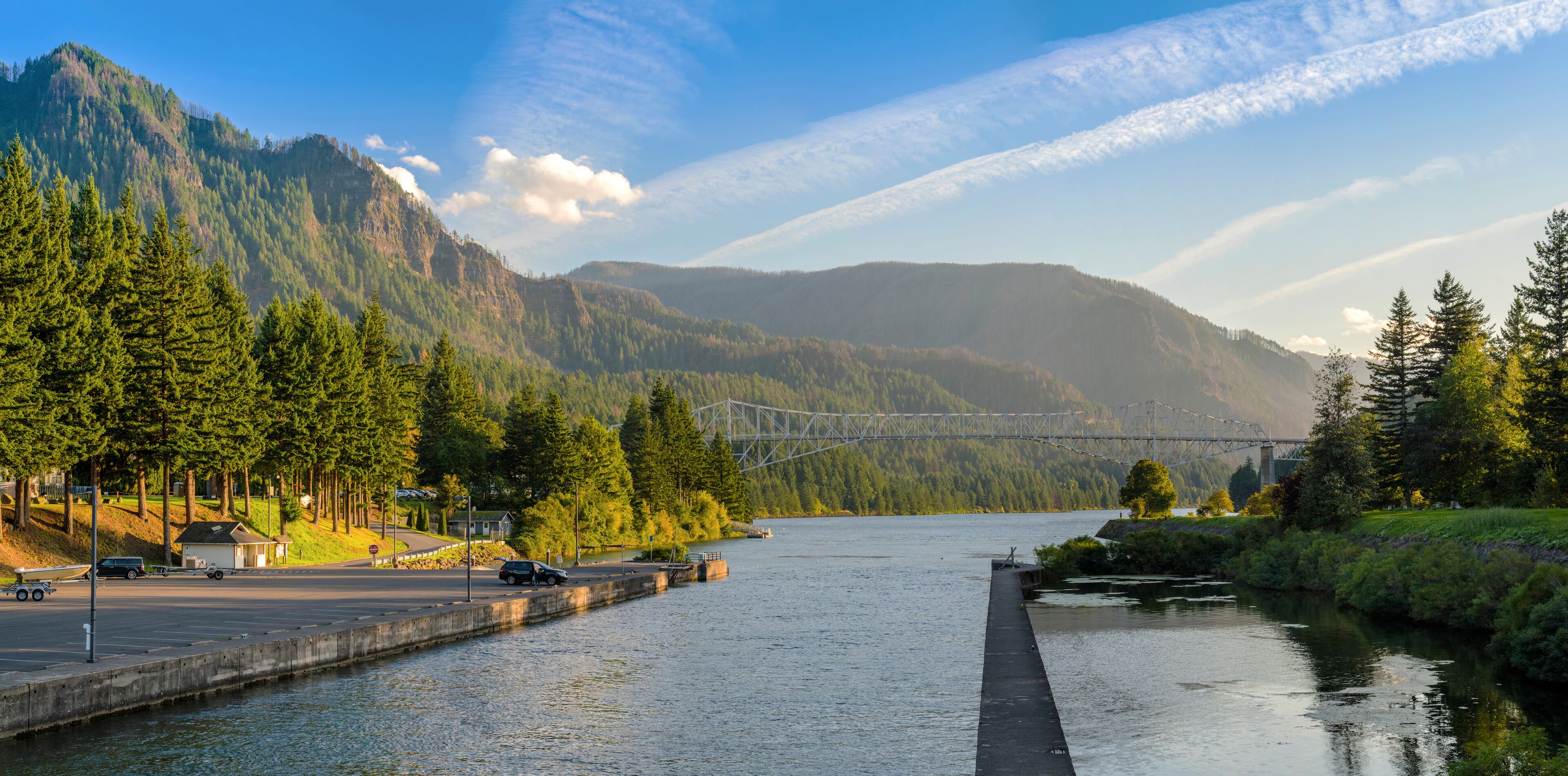 A river with a bridge, mountains, and a parking area with vehicles.