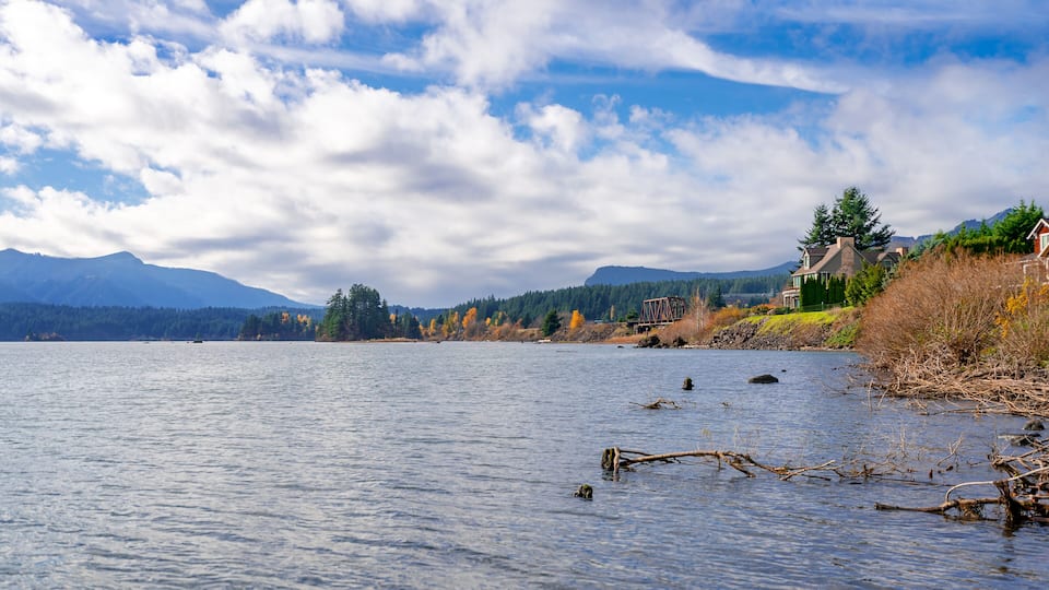 A calm lake with a wooden dock extending into the water, surrounded by autumn-colored trees and a mountain range in the background.
