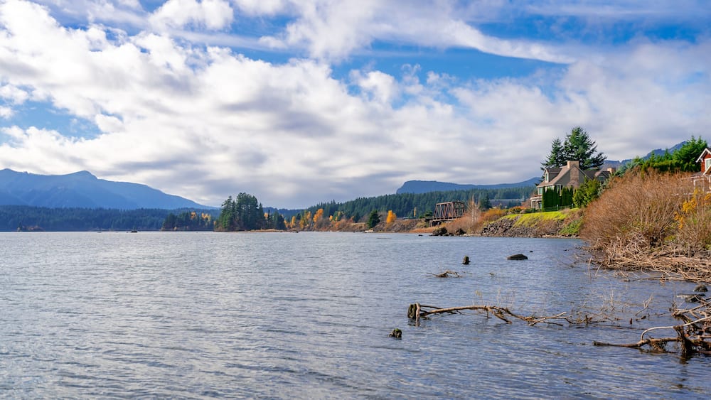 A calm lake with a wooden dock extending into the water, surrounded by autumn-colored trees and a mountain range in the background.