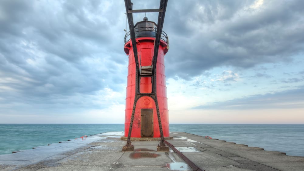 A red lighthouse on a pier with a cloudy sky.