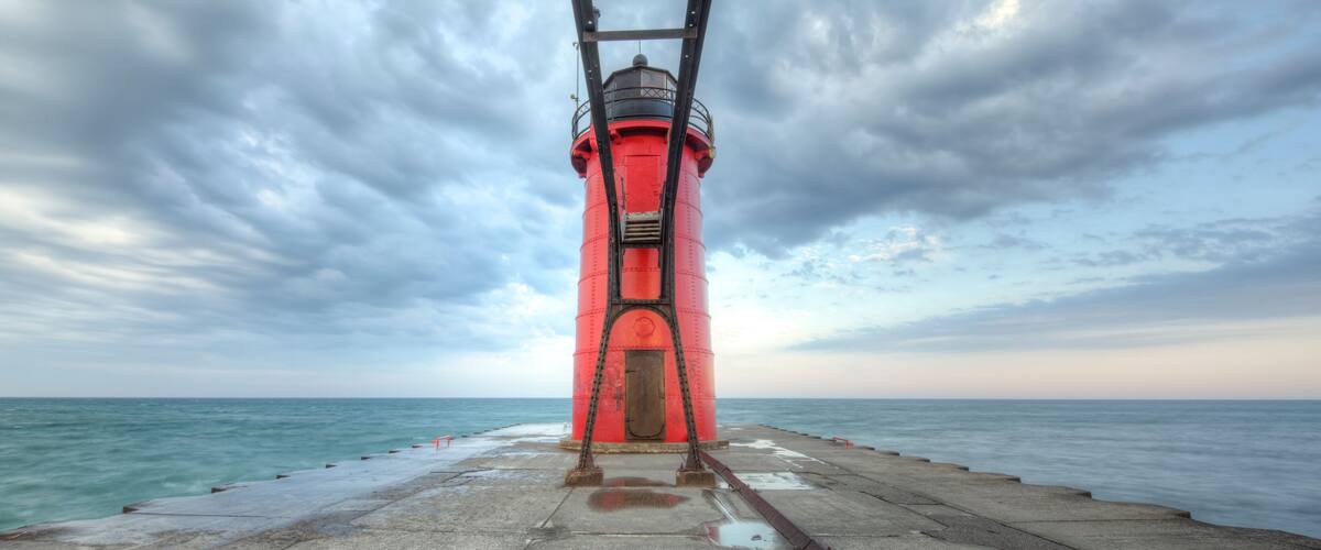 A red lighthouse on a pier with a cloudy sky.
