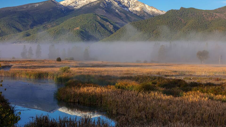 A misty mountain landscape with a reflective water body and dry grass fields.