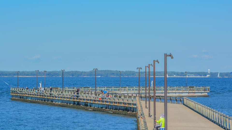 A pier extending into a body of water with people fishing, a clear sky, and distant land with a bridge.