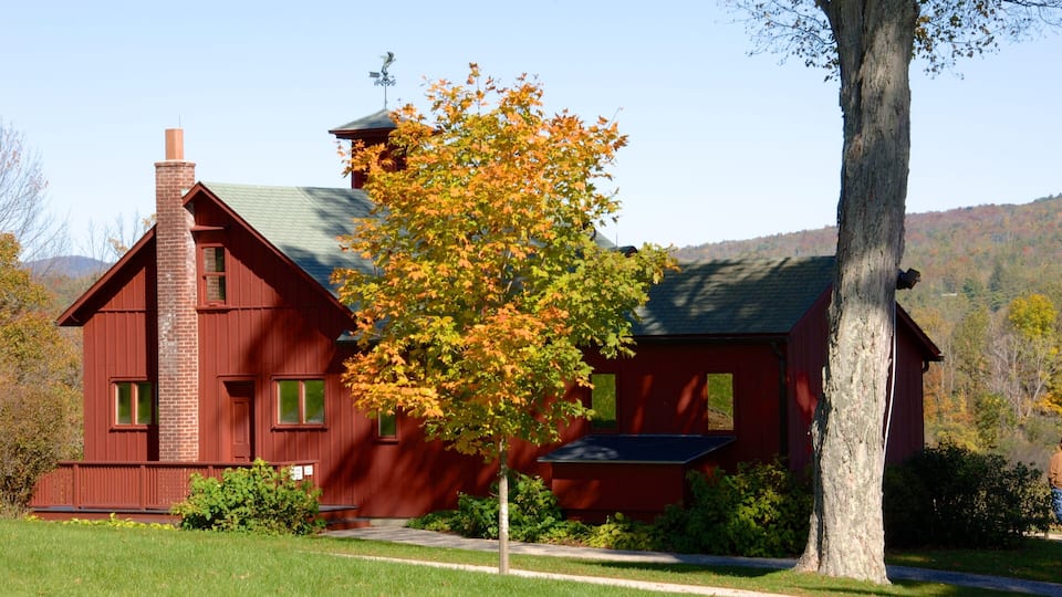 A red wooden house with a green roof, surrounded by a grassy yard and trees with autumn leaves.