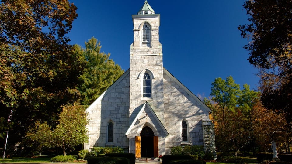 A stone church with a tall steeple and a cross on top, surrounded by trees and a well-maintained lawn.