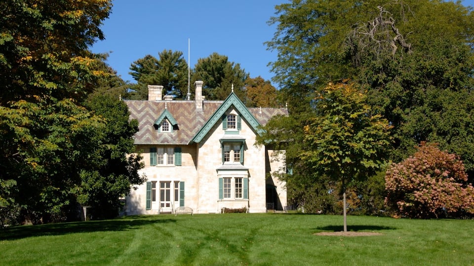 A large stone house with green shutters and a steep roof, surrounded by lush trees and a well-maintained lawn.