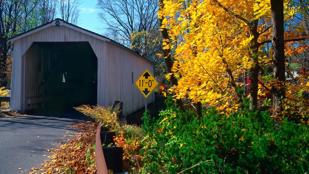 Eine gedeckte Brücke mit Höhenbeschränkungszeichen, umgeben von Herbstlaub.
