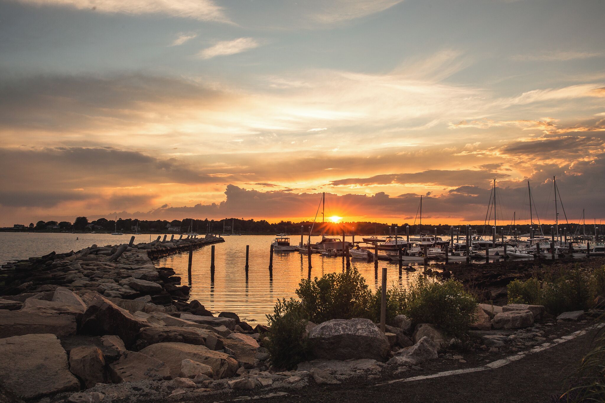 A marina at sunset with boats docked and a rocky shoreline.
