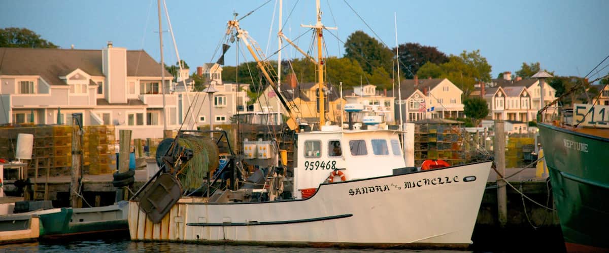 A fishing boat named 'SANDRA & MISCELLE' docked at a marina with houses and cranes in the background.