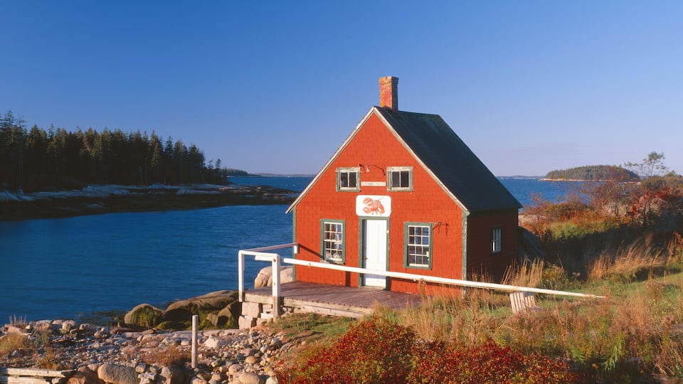 A red house by a rocky shore with a view of the ocean and a forested island in the distance.