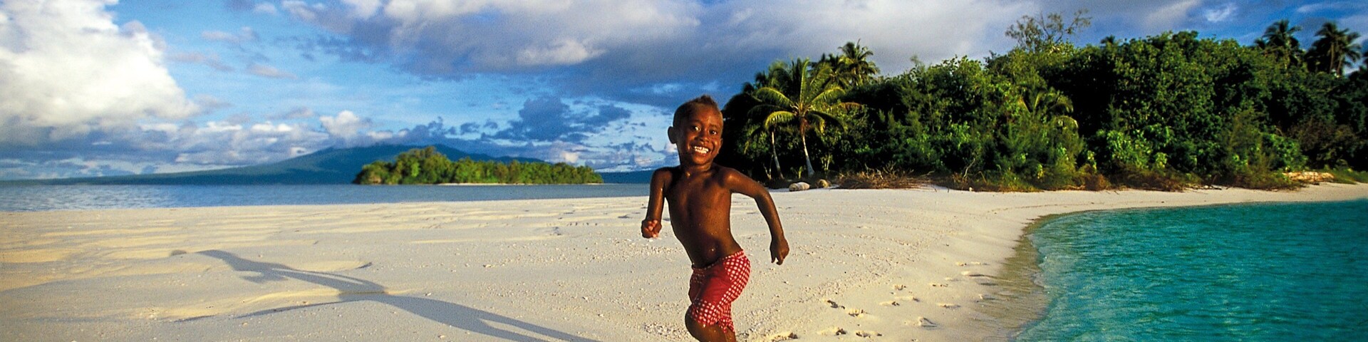 Niño corriendo en una playa arenosa con agua turquesa cristalina, palmeras y una montaña distante.